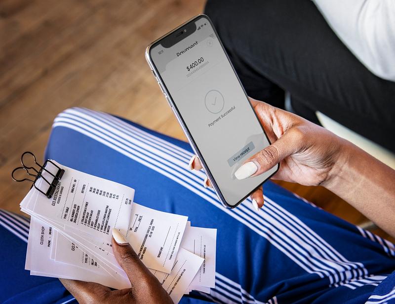 A woman doing banking and making payments on the smartphone she's holding in one hand, while also holding receipts in the other hand