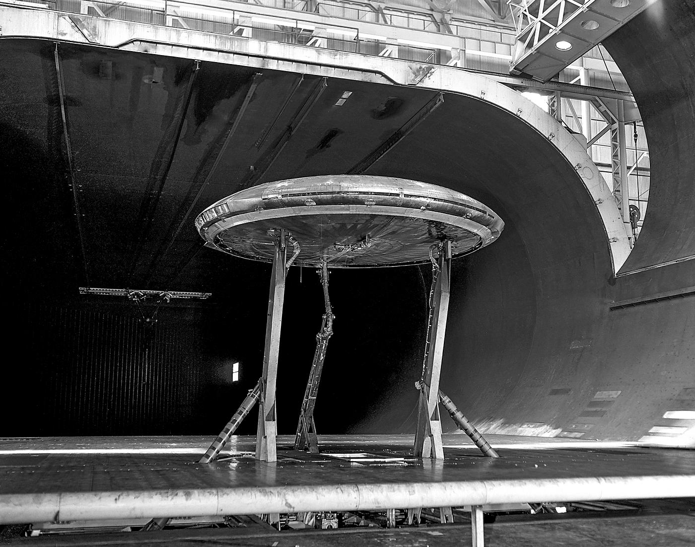 Avrocar Test in Ames 40x80 Foot Wind Tunnel. Rear view of the Avrocar with tail, mounted on variable height struts. Overhead... Avrocar Test in Ames 40x80 Foot Wind Tunnel. Rear view of the Avrocar with tail, mounted on variable height struts. Overhead...